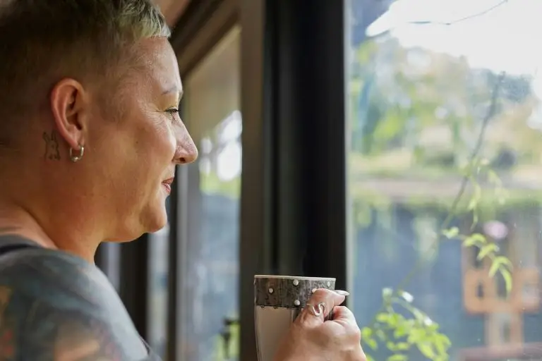 Woman looking out of a window into the garden, cup of coffee in hand